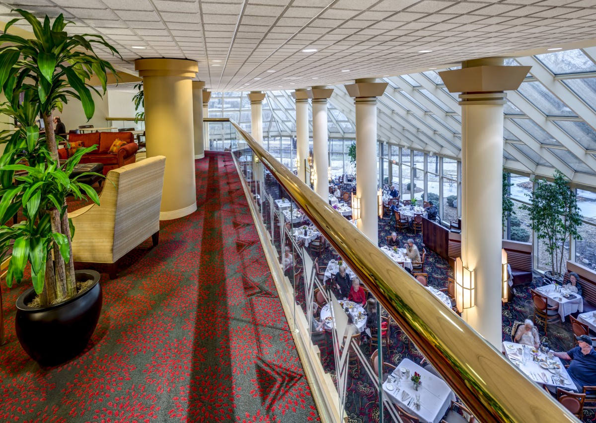 A spacious restaurant interior featuring a balcony view overlooking a dining area filled with neatly arranged tables and chairs, supported by large columns and a patterned carpet floor.