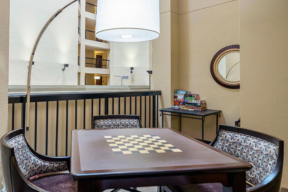 A cozy indoor setting featuring a checkers game table, surrounded by chairs and illuminated by a large lamp.
