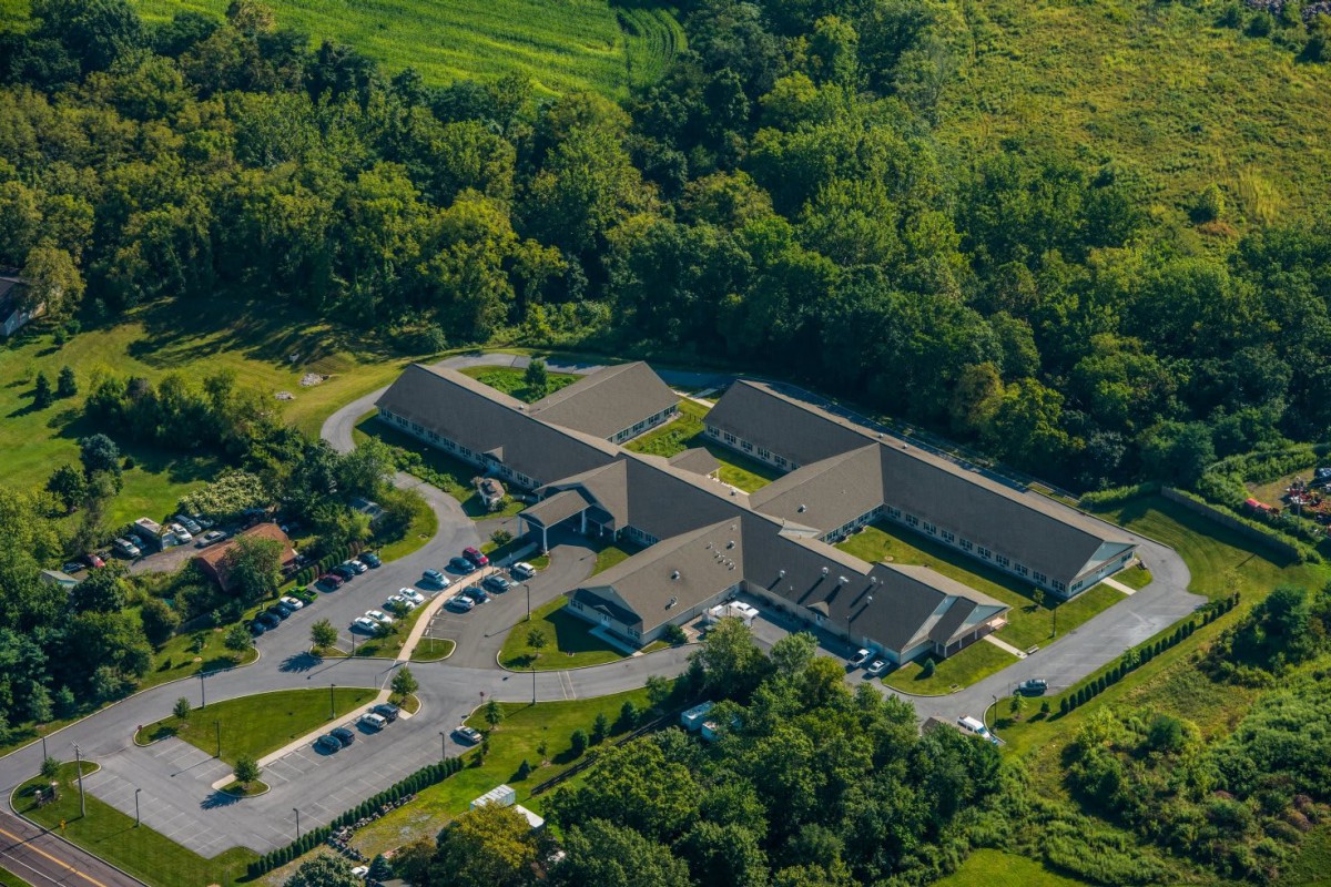 Aerial view of a complex of buildings surrounded by lush greenery and a parking area.