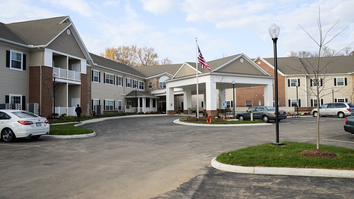 The image shows a large building with an entrance driveway and several parked cars, featuring a flagpole with an American flag.
