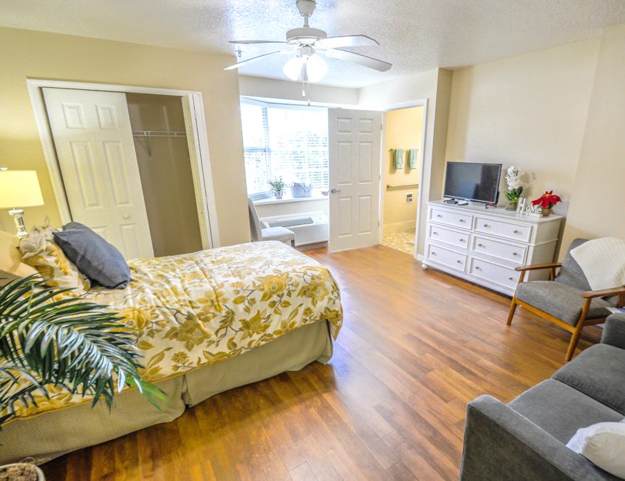 A well-lit bedroom with a bed, dresser, and TV on a wooden floor.