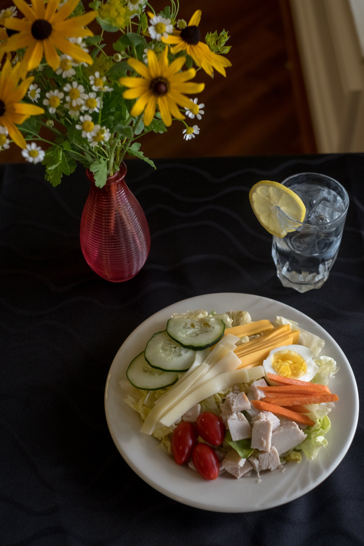 A healthy salad with chicken, boiled egg, and vegetables accompanied by a glass of lemon water and a vase of fresh flowers.