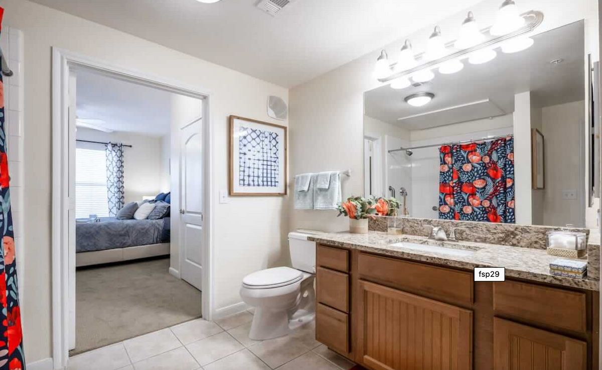 A modern bathroom with a view into the adjacent bedroom, featuring a large vanity, well-lit mirror, and contemporary decor.