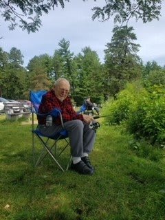 An elderly person sitting on a camping chair, fishing by a lakeside surrounded by lush greenery.