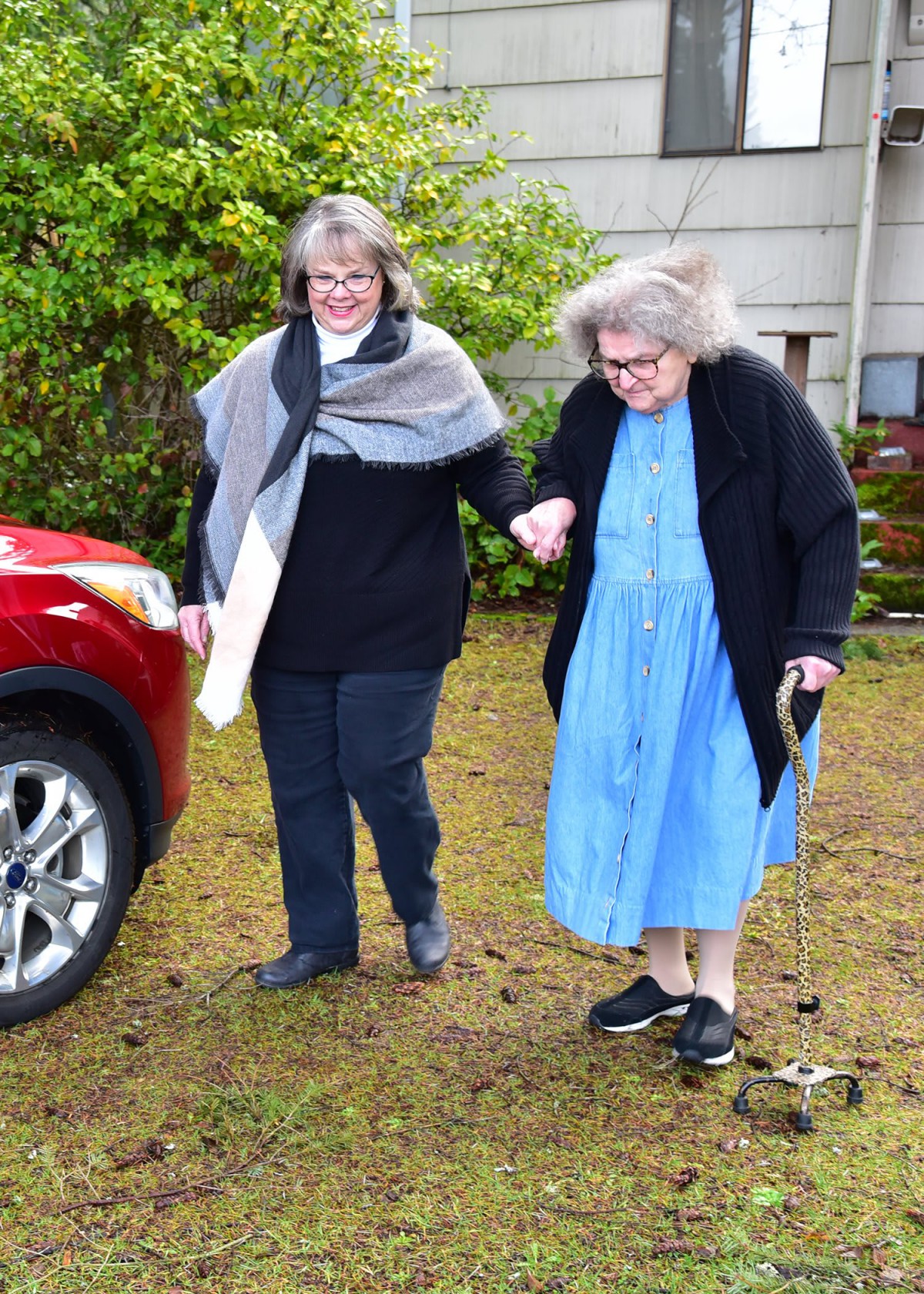 An elderly woman is assisted by another person as they walk together outside near a car.