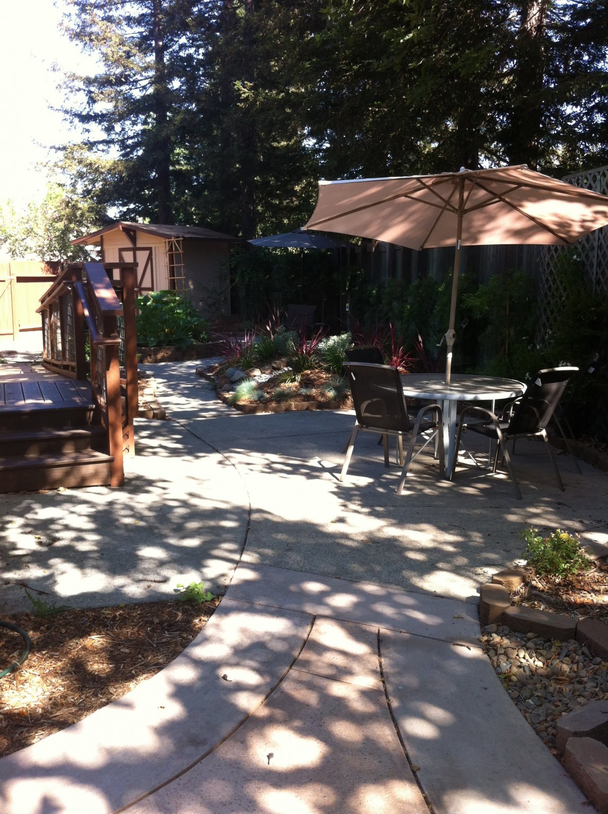A tranquil backyard patio with outdoor furniture and an umbrella shaded by tall trees.