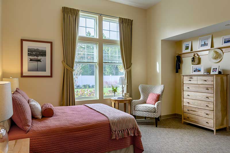 A cozy bedroom filled with natural light, featuring a bed with a pink coverlet, an armchair by the window, and a wooden dresser.