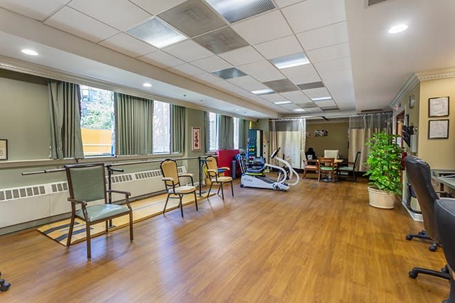 A spacious therapy room with exercise equipment and seating, featuring large windows and bright lighting.