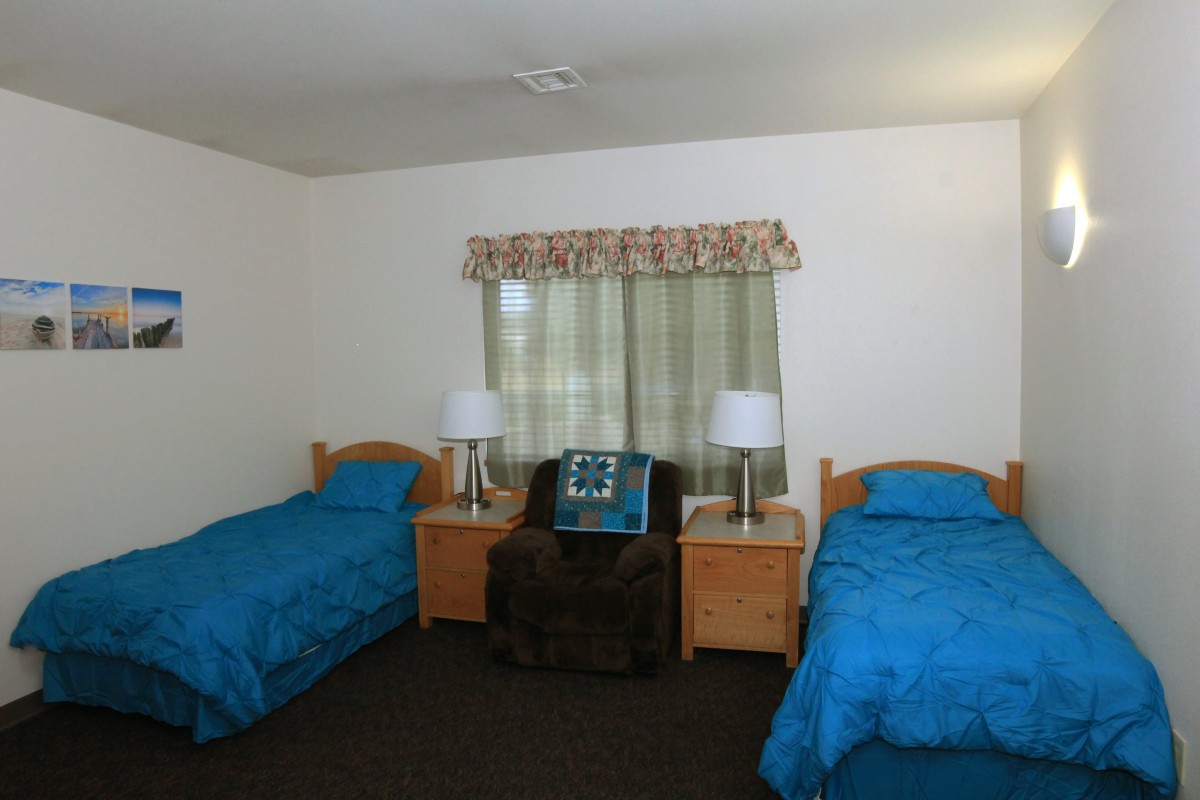A simple bedroom with two twin beds and blue bedding, featuring a nightstand between the beds and a window with patterned curtains.