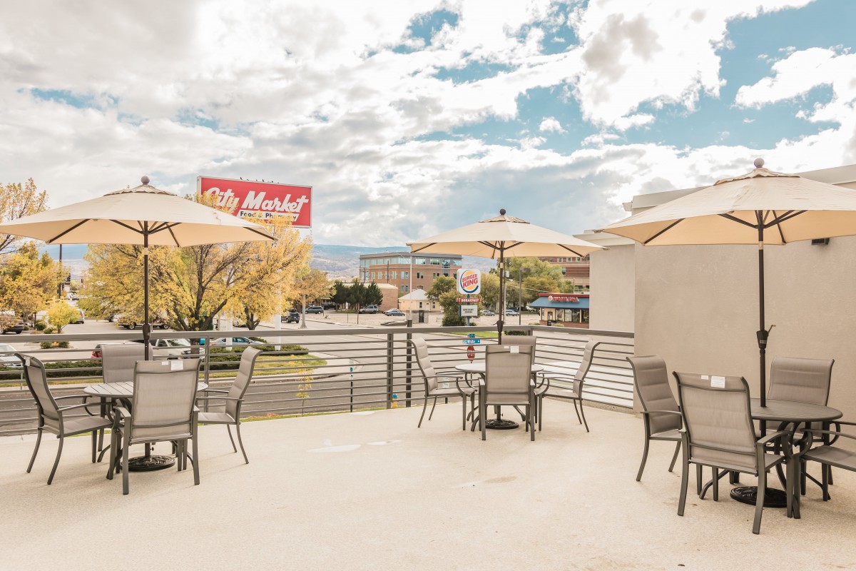 A patio with tables and chairs under umbrellas, set against a city backdrop under a cloudy sky.