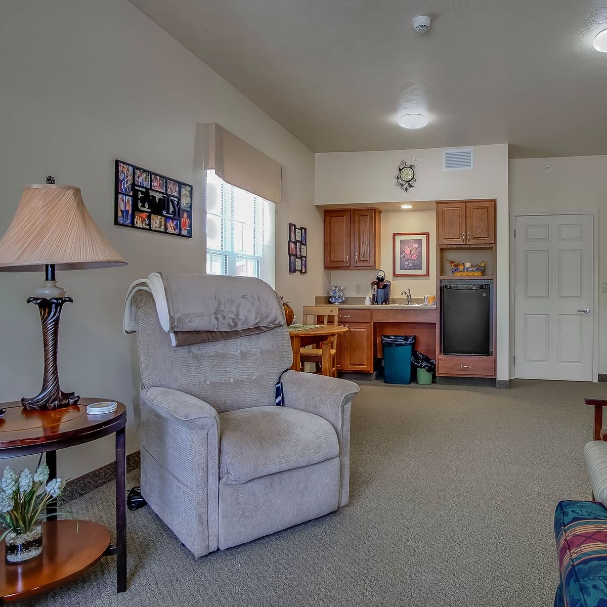 A cozy living room area with a comfortable armchair, side table, and a view of a kitchenette in the background.