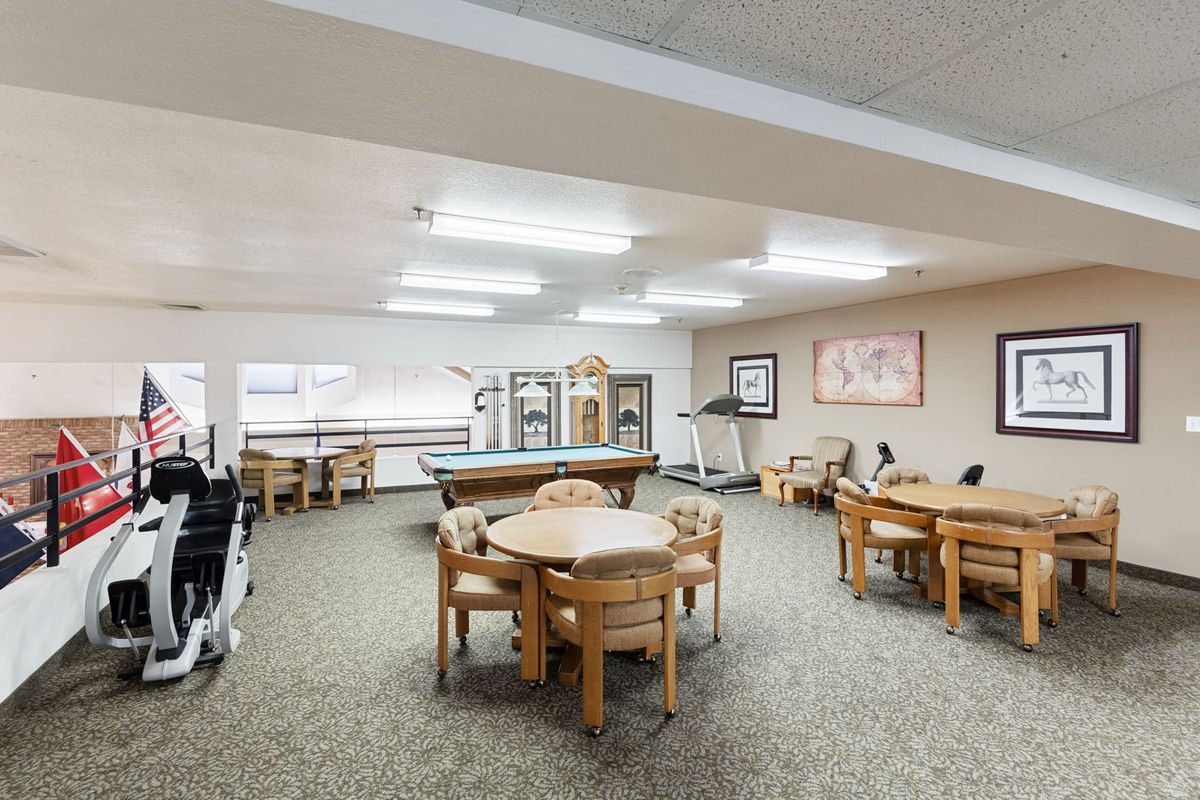 A spacious game room featuring multiple tables, chairs, a pool table, treadmill, and various exercise equipment.