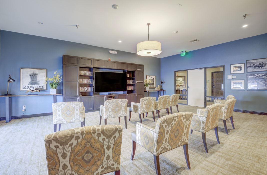 A modern meeting room with patterned chairs arranged facing a large television, surrounded by bookshelves and stylish lighting.