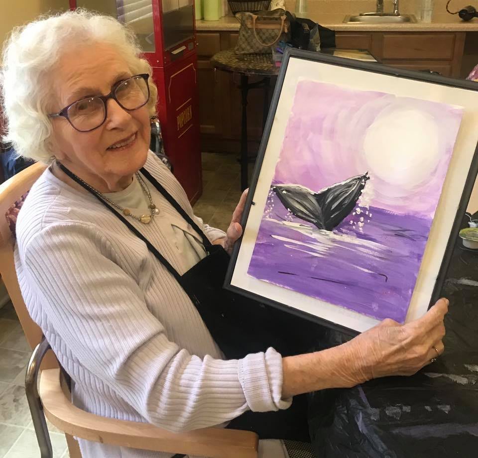 An elderly woman proudly displays her painting of a whale against a purple ocean backdrop.