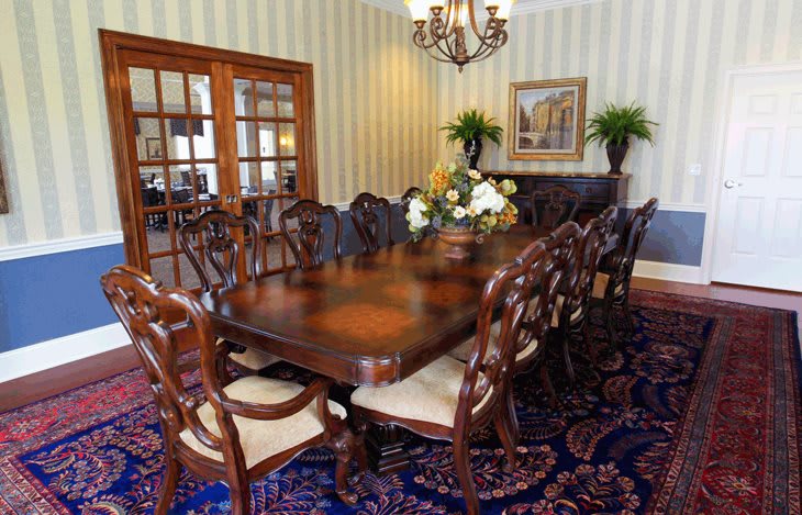 A traditional dining room featuring a large wooden table surrounded by ornate chairs, a chandelier overhead, and floral decorations.