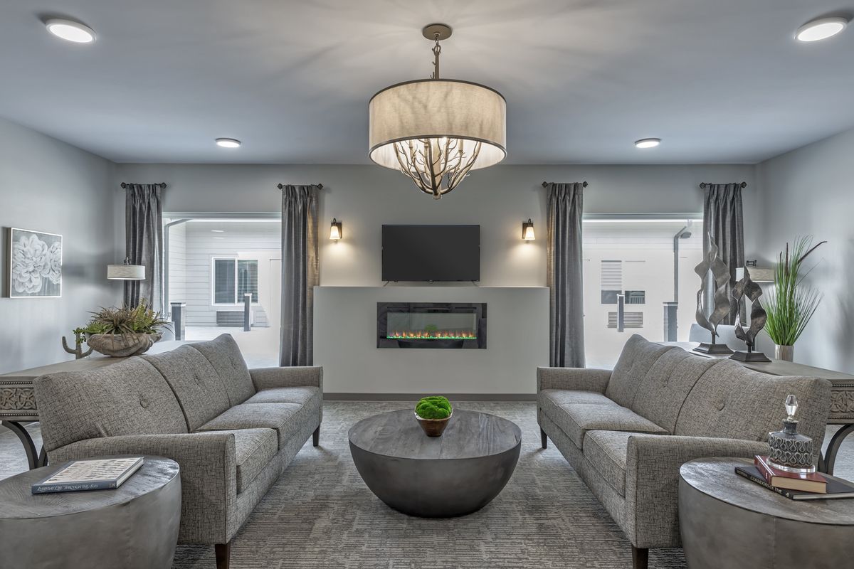 A modern living room featuring gray sofas, a central coffee table, a chandelier, and a cozy fireplace with a flat-screen TV above.