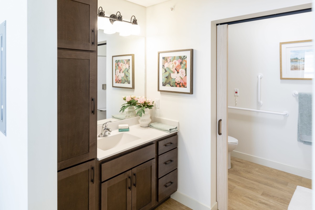 A modern bathroom with a wooden cabinet, artwork on the wall, and a vase of flowers on the counter.