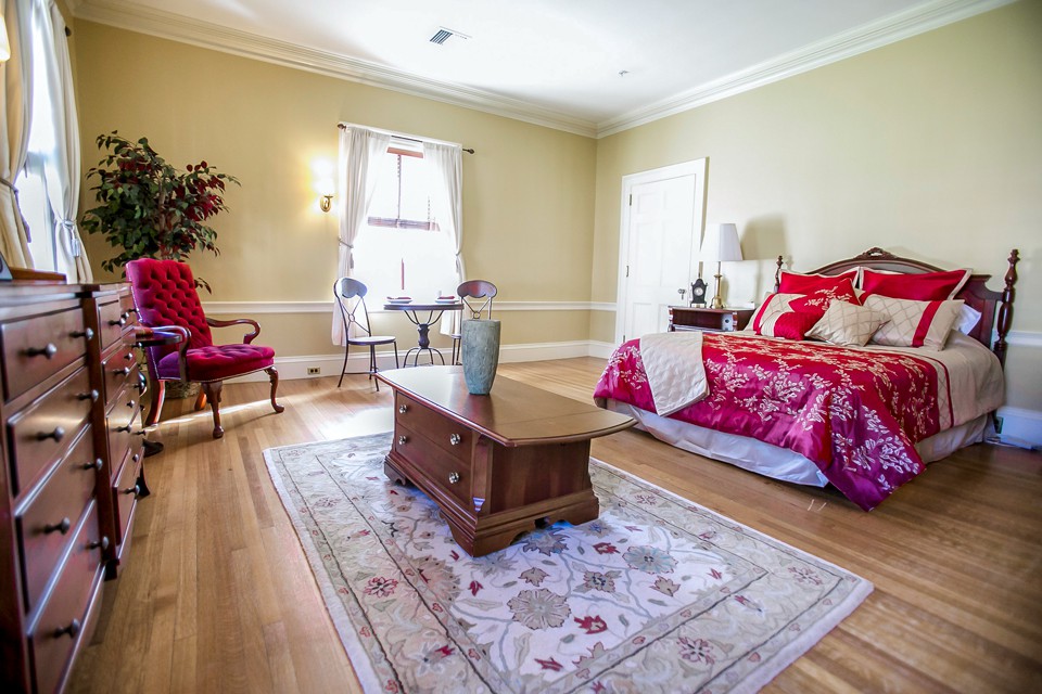 A cozy bedroom with red bedding, wooden floors, and natural light streaming in.