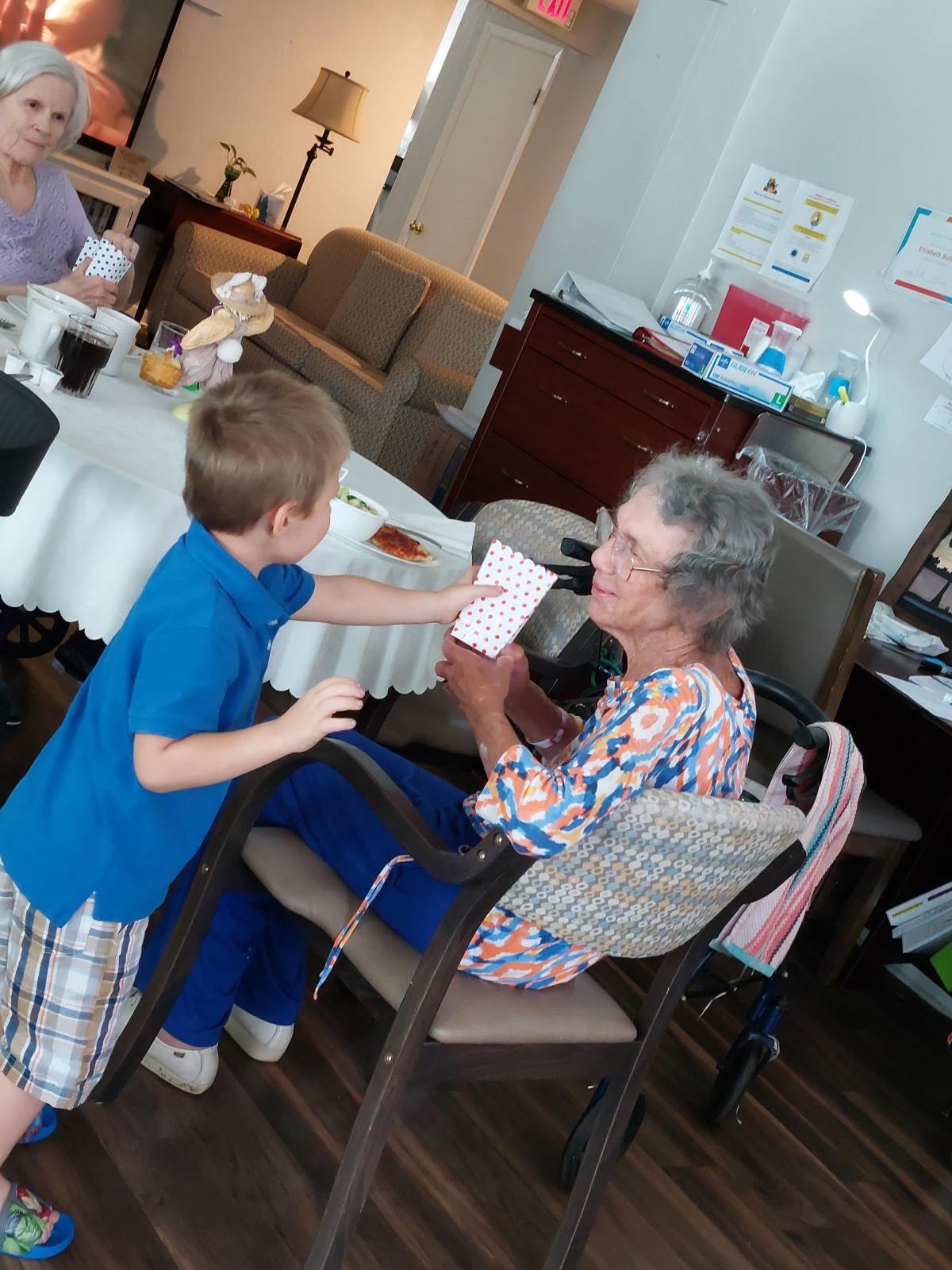 A child handing a card to an elderly woman in a cozy room setting.