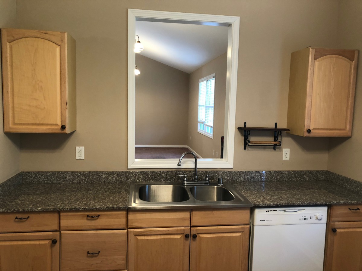 A kitchen with wooden cabinets, a granite countertop, and a window overlooking another room.