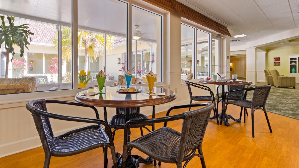 A cozy dining area with tables and chairs, featuring bright indoor lighting and large windows.