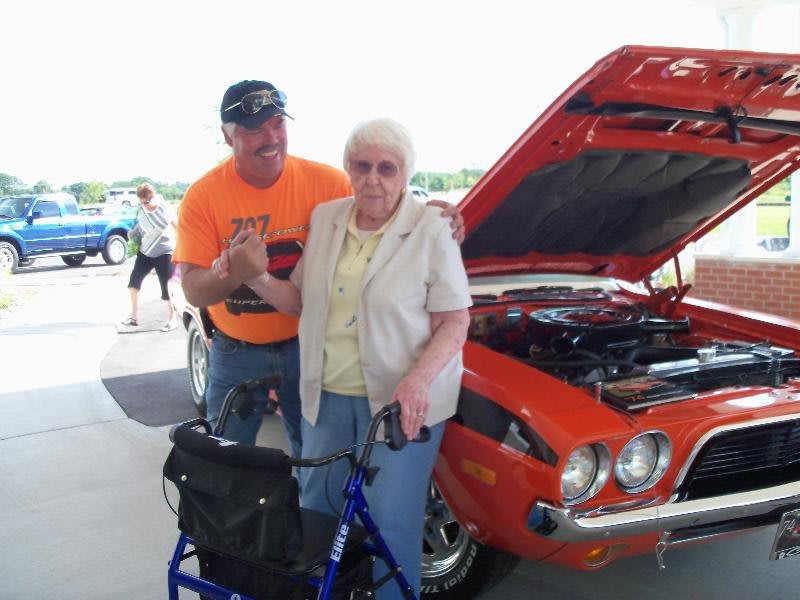 An elderly woman with a walker stands beside a man in front of an orange classic car at a car show.