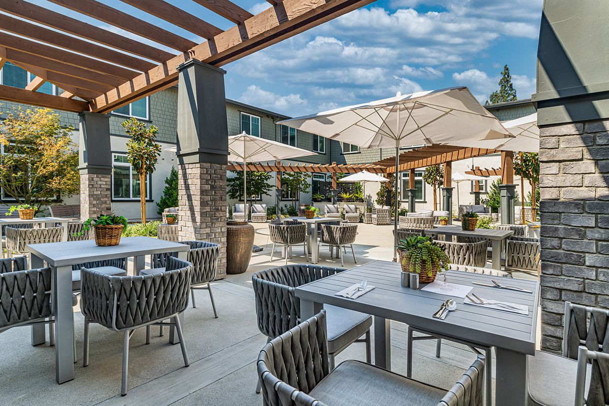 A modern outdoor dining area with tables, chairs, and umbrellas under a pergola.