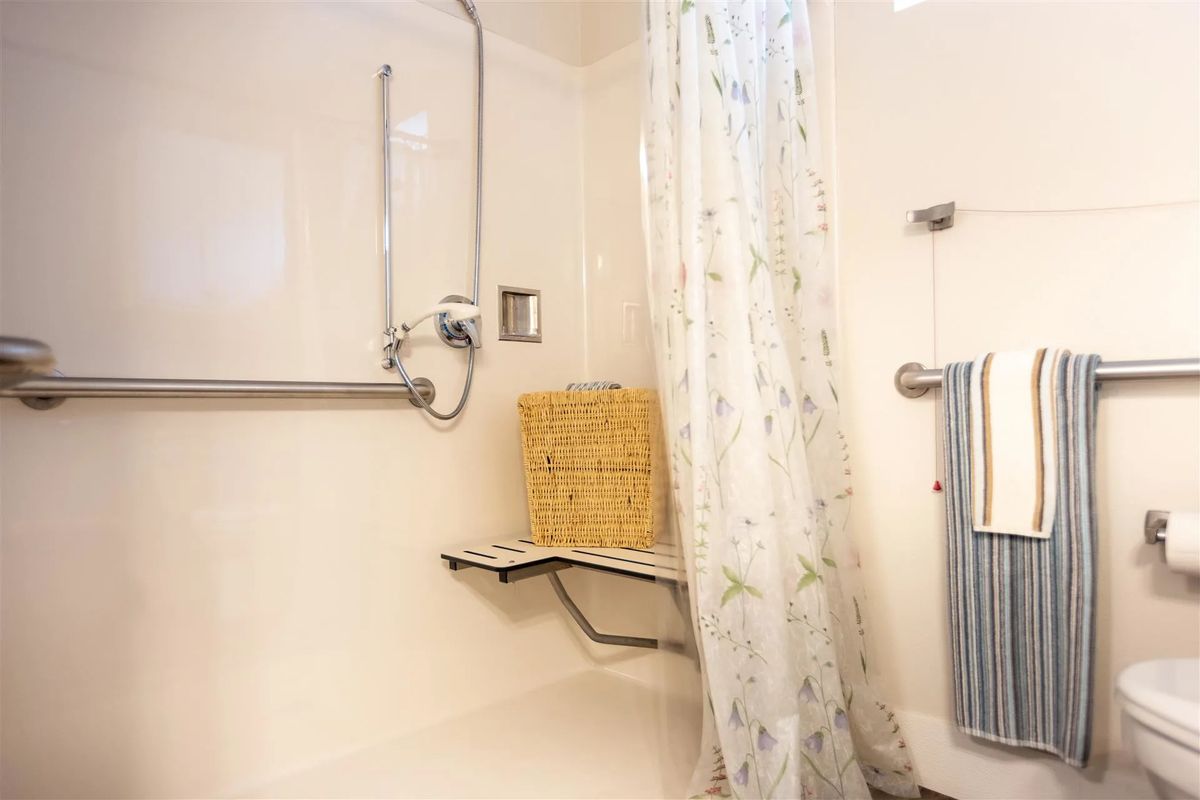 An accessible bathroom with a shower seat and grab bar, featuring a floral shower curtain and towel rack.