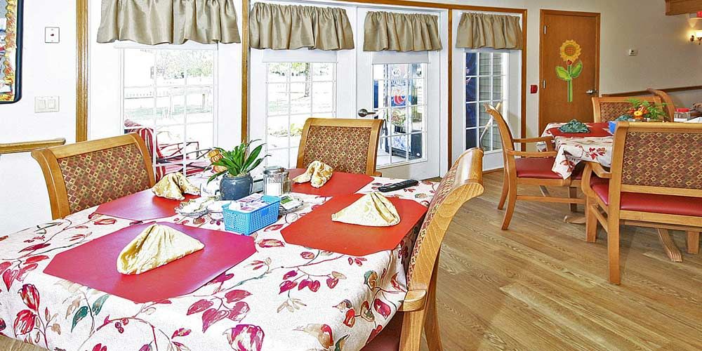 A bright dining room with a floral tablecloth, neatly arranged place settings, and large windows letting in natural light.