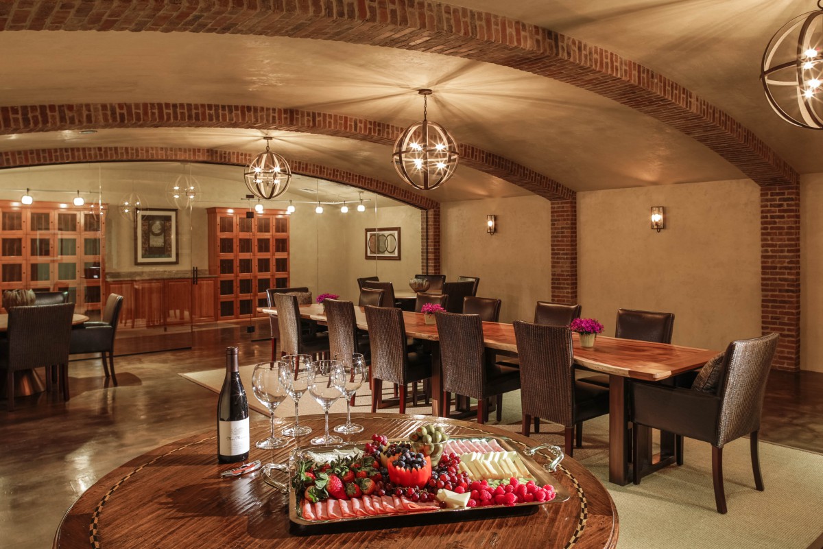 A luxurious dining area set in a wine cellar with a large table, elegantly arranged glasses, and a food platter.