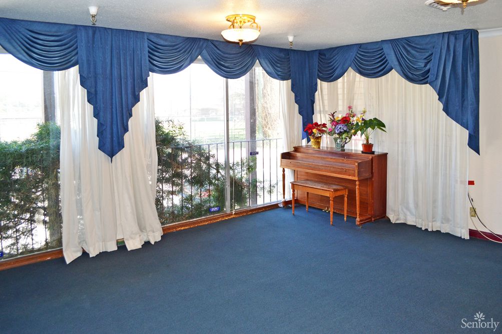 A living room featuring a piano, large windows with blue and white draped curtains, and a blue carpeted floor.