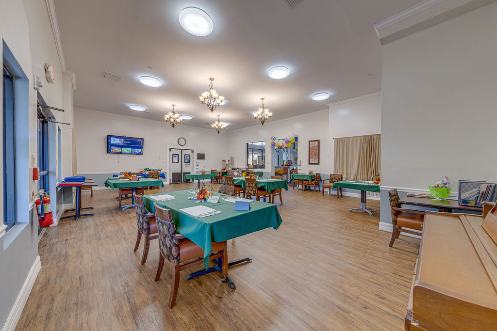 A spacious dining room with tables covered in green tablecloths, surrounded by chairs and illuminated by ceiling lights.