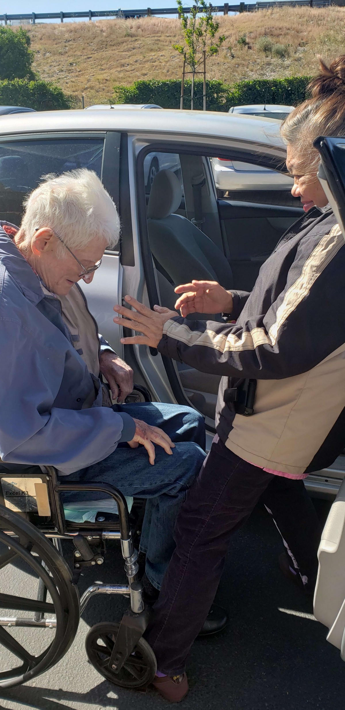 A caregiver assisting an elderly person in a wheelchair by a car.