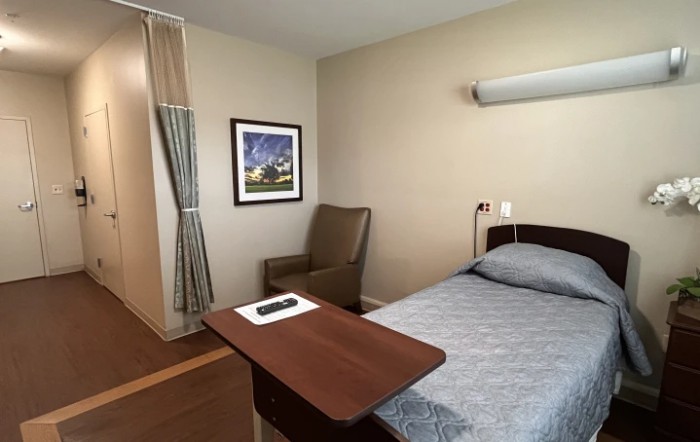 A clean and simple hospital room with a single bed, chair, and artwork on the wall.