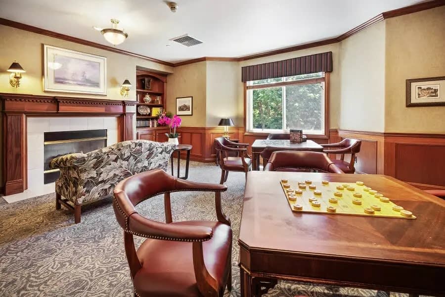 A cozy living room with leather chairs, a wooden table set for checkers, a fireplace, and natural light streaming through a large window.