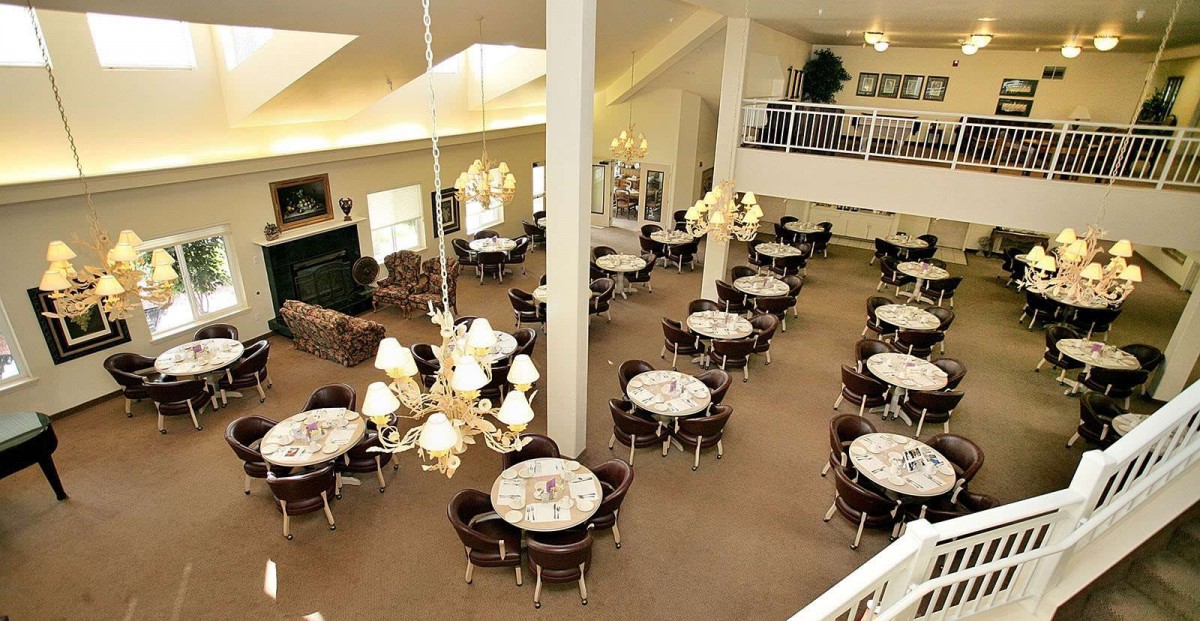 A spacious dining area with multiple round tables set with white tablecloths, elegant chandeliers, and a carpeted floor.