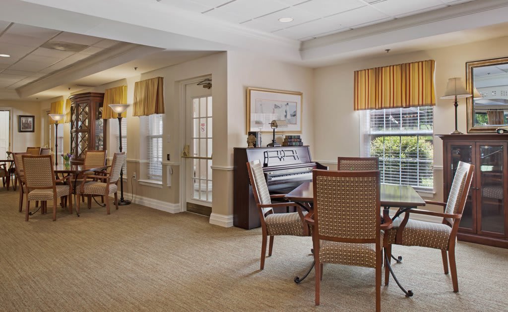 A cozy living area featuring a piano, seating arrangement with chairs and tables, and large windows with blinds.