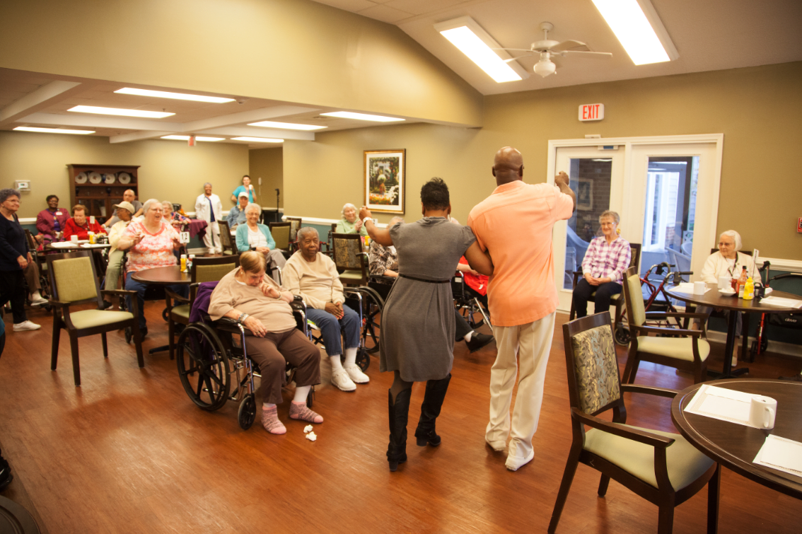 A group of elderly individuals gathered in a care facility, with some participating in a dance activity.