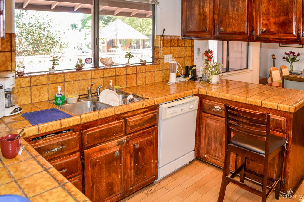 A cozy kitchen with wooden cabinets and tile countertops, featuring a dishwasher and a window with a view.