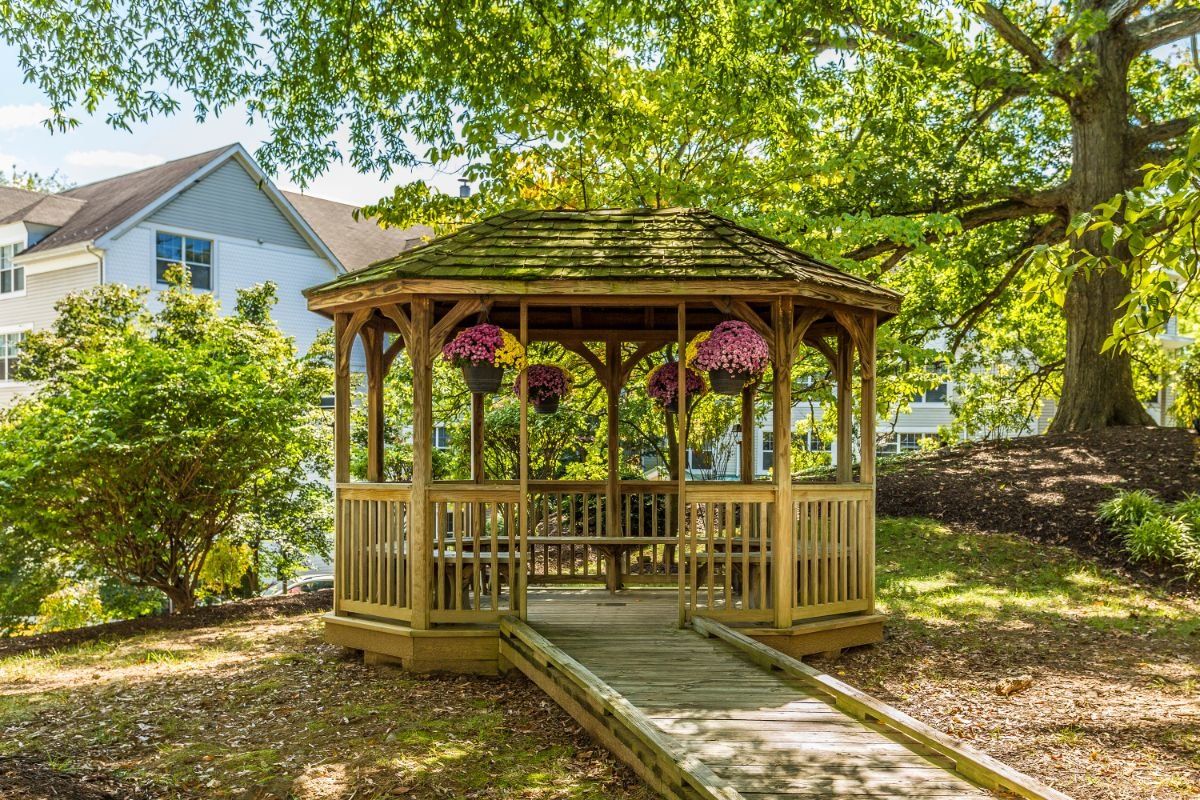 A wooden gazebo in a garden setting, adorned with hanging plants and surrounded by trees.