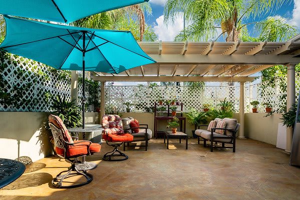 A cozy outdoor patio with comfortable seating and a vibrant umbrella under a pergola surrounded by lush greenery.