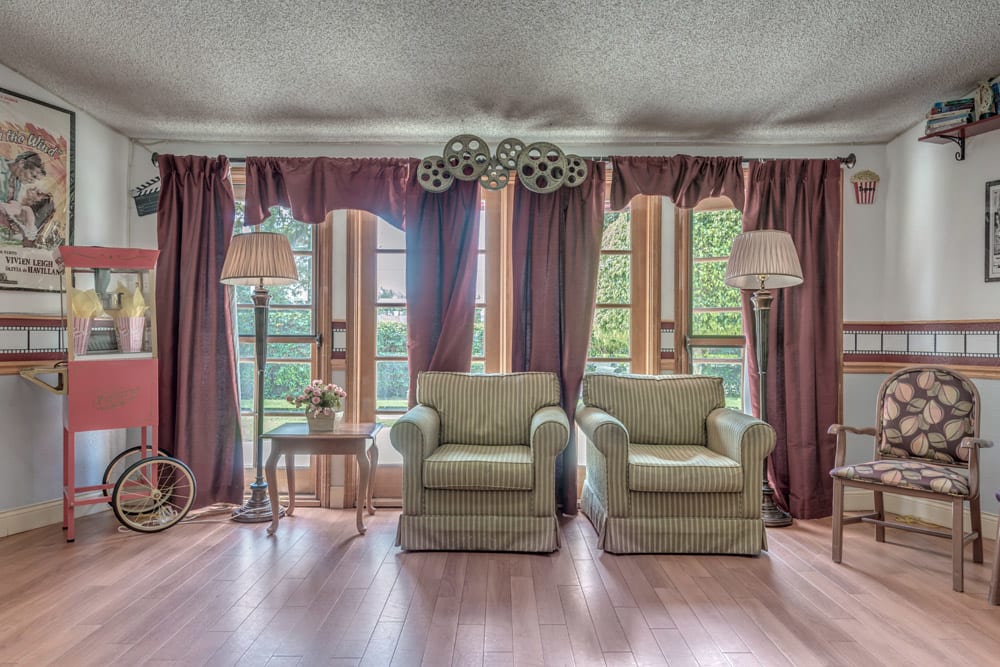 A cozy living room with two armchairs, flanked by elegant curtains and standing lamps, overlooks a lush garden through large windows.