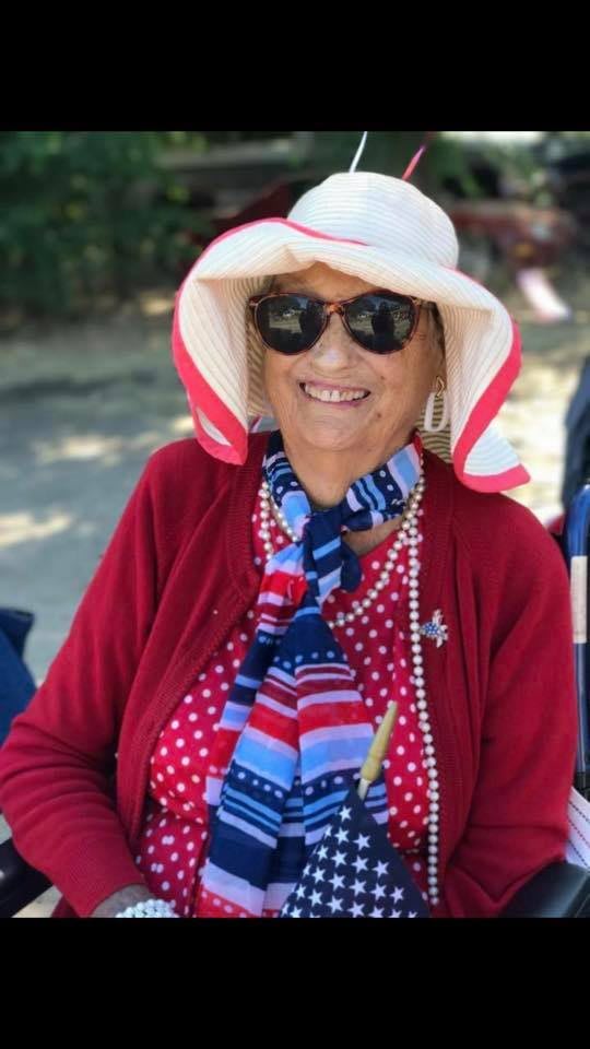 A smiling woman wearing sunglasses, a sun hat, and a colorful scarf sits outdoors.