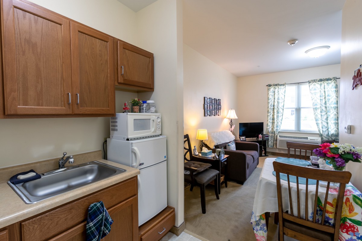 A cozy apartment with an open kitchen and living room layout, featuring wooden cabinets, a microwave, a dining table, and comfortable seating.