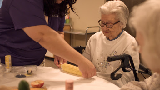 A caregiver assists an elderly woman with a cooking activity.