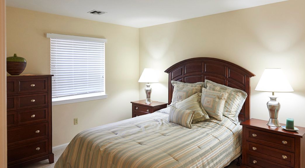 A neatly arranged bedroom with a bed, pillows, and a nightstand with a lamp by the window.