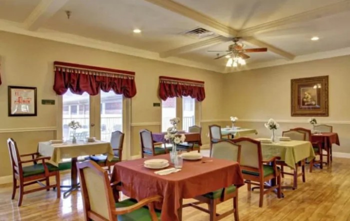 A cozy dining area with tables set for meals, featuring large windows with red curtains and a ceiling fan overhead.