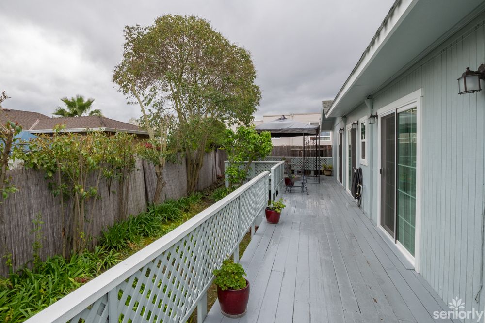A long wooden deck with potted plants, surrounded by a fence and trees.