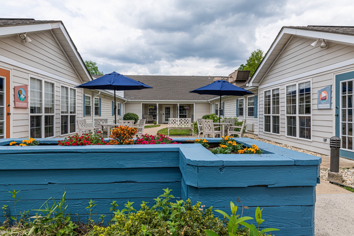 A residential courtyard featuring vibrant gardens with blue planters, outdoor seating, and umbrellas under a cloudy sky.