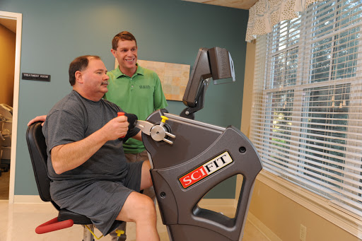 A man engages in a rehabilitation exercise with the assistance of a trainer, using specialized gym equipment.